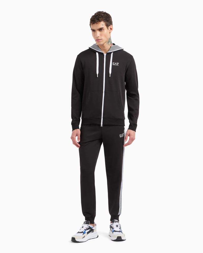 A man poses in a black EA7 tracksuit and white sneakers in a studio setting. He faces the camera with a neutral expression and stands still.