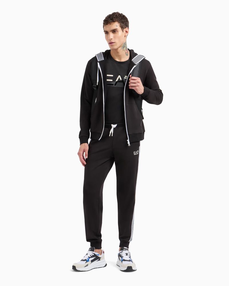 Man poses in black EA7 tracksuit with zip hoodie, tee, sweatpants, and white/blue sneakers against a white background in a studio shot.
