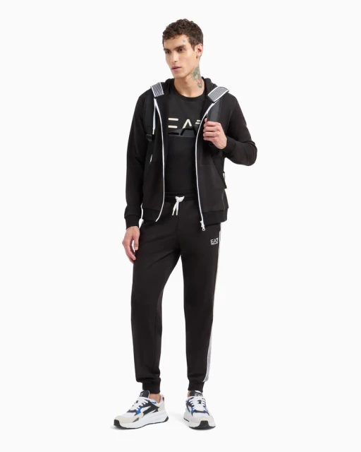 Man poses in black EA7 tracksuit with zip hoodie, tee, sweatpants, and white/blue sneakers against a white background in a studio shot.