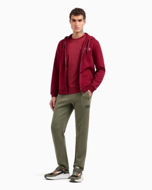 A man stands in a photo studio wearing a red hooded sweatshirt, red t-shirt, olive green sweatpants, and multi-colored sneakers against a white background.