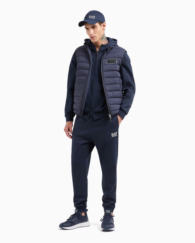 A man in a navy hooded tracksuit and vest, standing in a photo studio against a white backdrop, wearing matching shoes and baseball cap.