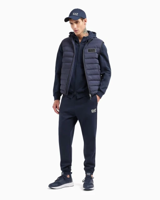 A man in a navy hooded tracksuit and vest, standing in a photo studio against a white backdrop, wearing matching shoes and baseball cap.