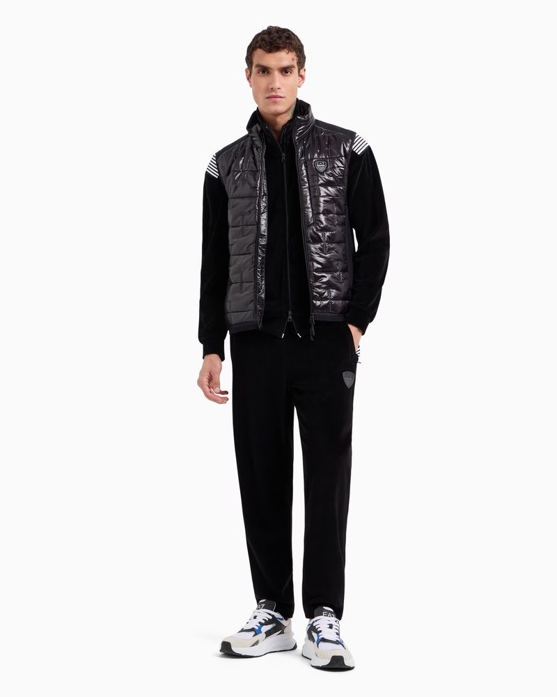 A full-shot studio image shows a male model wearing a quilted vest, velvet tracksuit, and sneakers against a bright white backdrop.