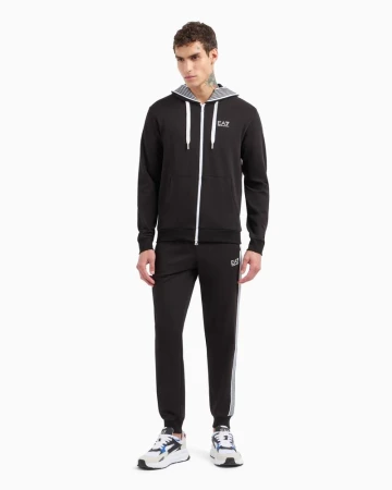 Full-length studio portrait of a male model wearing a black zip-up tracksuit and sneakers against a plain white background.