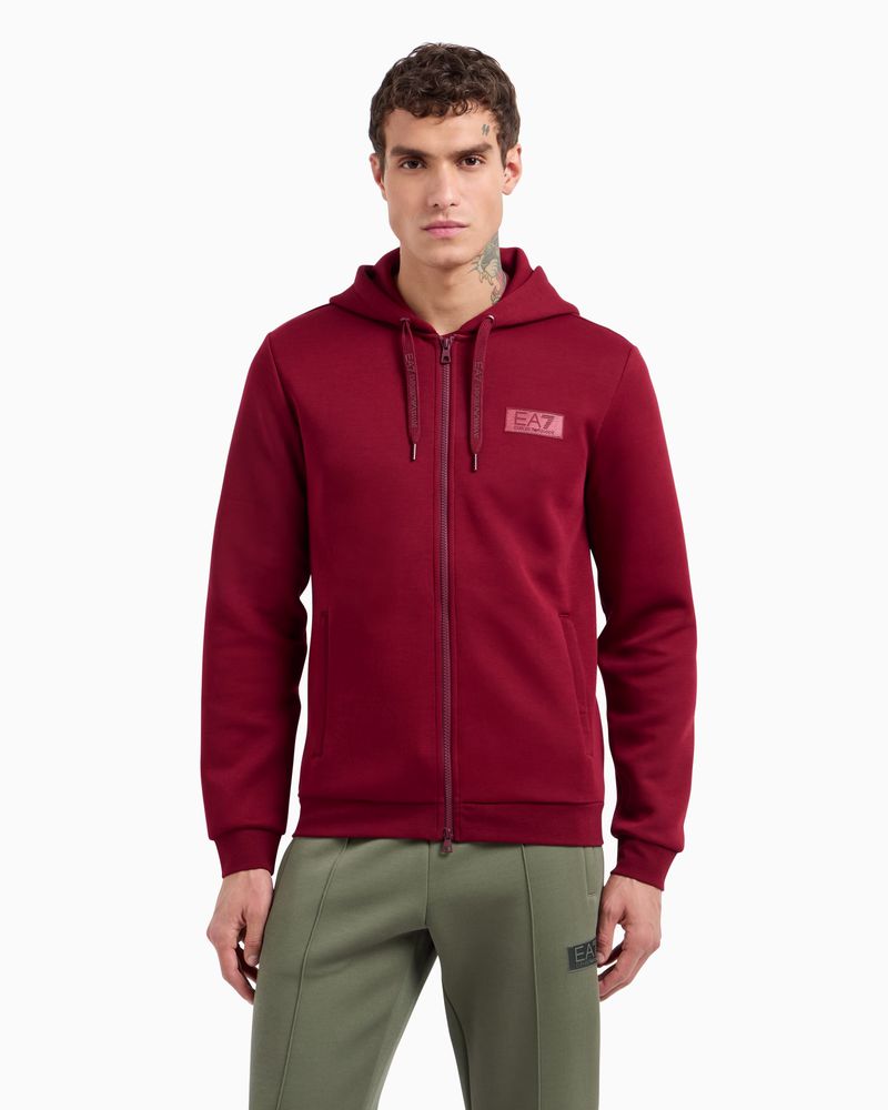 Male model wearing a burgundy zip-up hoodie and green sweatpants against a white background for Emporio Armani EA7