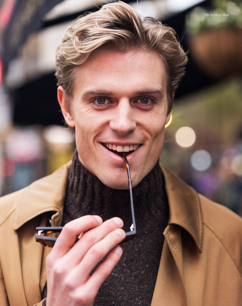Smiling man with blond hair in a brown turtleneck and tan coat holding glasses with outdoor background, portrait shot, fashion.