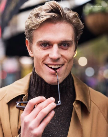 Smiling man with blond hair in a brown turtleneck and tan coat holding glasses with outdoor background, portrait shot, fashion.