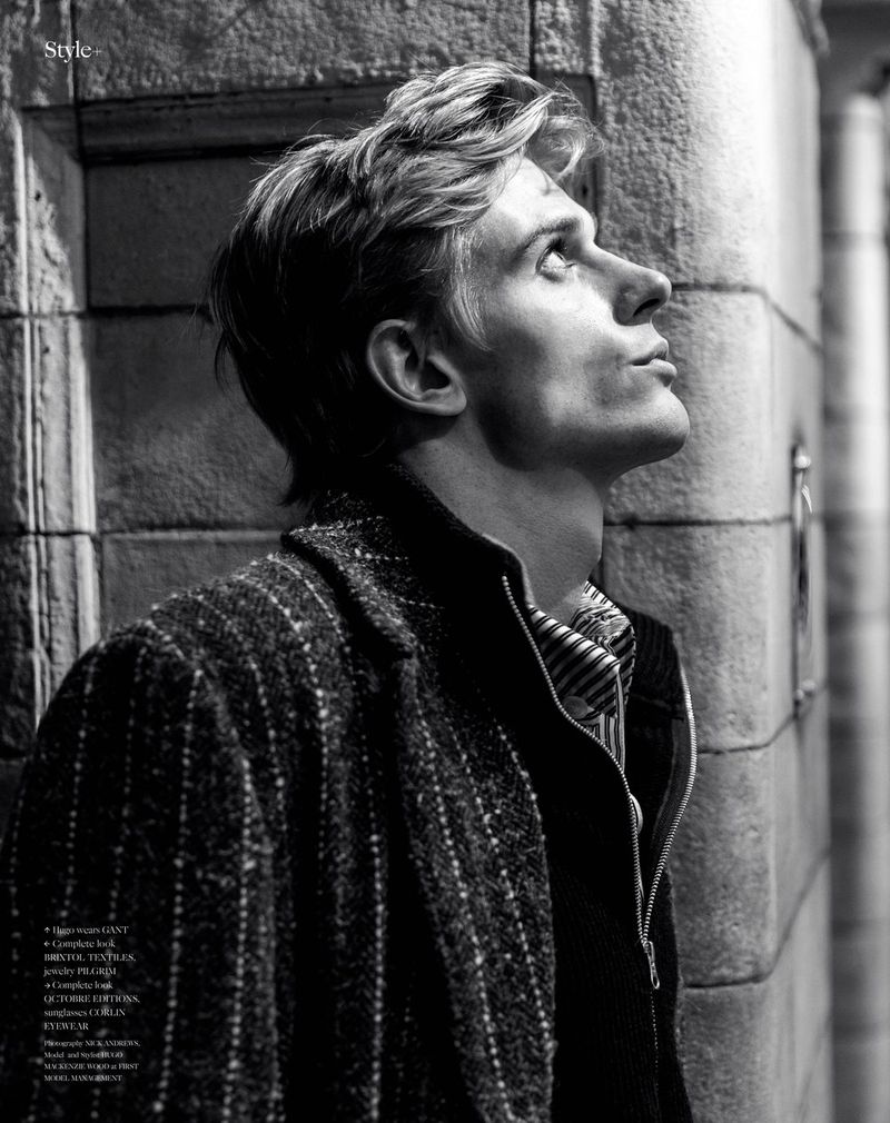 Black and white portrait of a fair-skinned man wearing a jacket and collared shirt, looking up next to a textured brick wall.