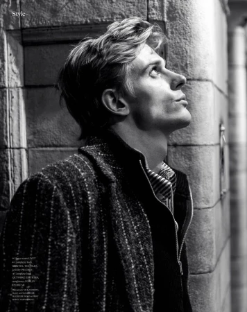 Black and white portrait of a fair-skinned man wearing a jacket and collared shirt, looking up next to a textured brick wall.