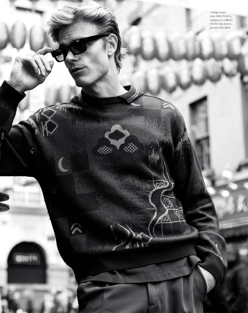 Monochrome portrait of a blonde male model wearing sunglasses and a patterned sweater in an outdoor urban setting.