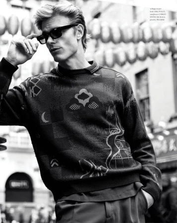 Monochrome portrait of a blonde male model wearing sunglasses and a patterned sweater in an outdoor urban setting.