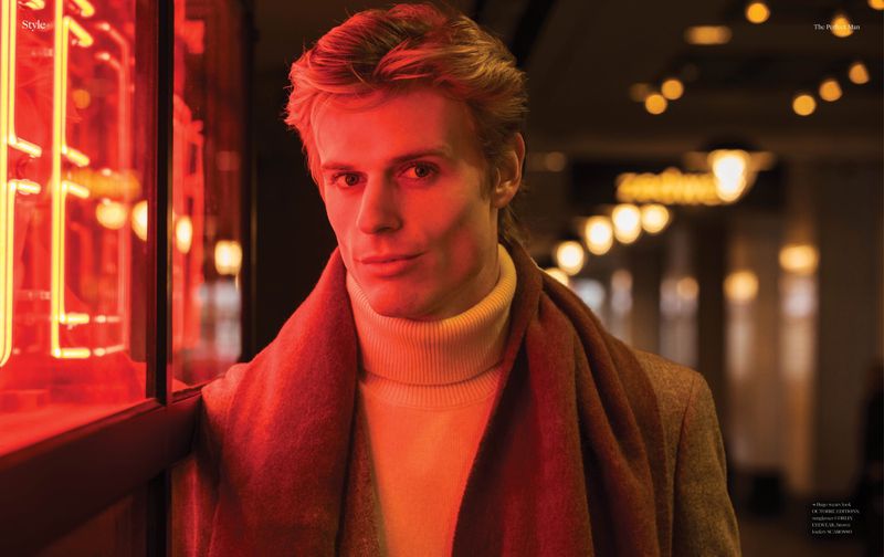 Portrait of a blonde-haired man in a turtleneck and coat, standing near a bright red neon sign with soft focus in background.
