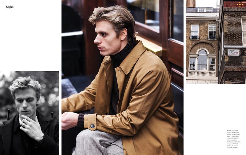 Male model in a tan trench coat poses in an urban setting; buildings visible in the background, showcasing men's fashion.