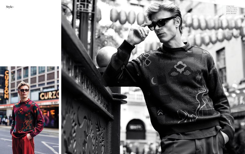 Monochrome portrait of a young adult male in sunglasses and patterned sweater leaning on a metal railing in an urban environment.