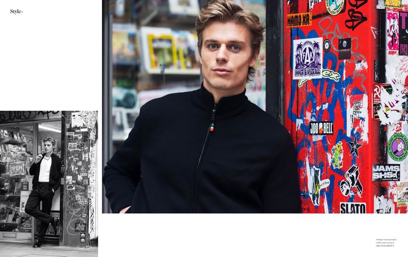 A fair-skinned man with blonde hair is leaning against a red, sticker-covered wall next to a magazine stand for a fashion editorial.