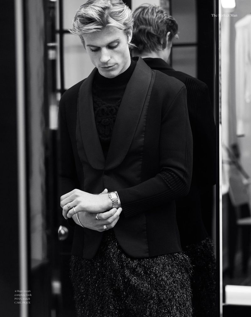 Monochrome portrait of a blonde male model wearing a black jacket and patterned top, indoors with a mirror in the background.