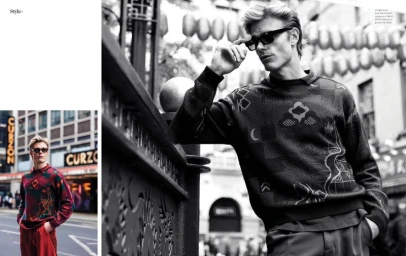 Black and white editorial portrait of a man in sunglasses wearing a patterned sweater leaning against a railing on a city street