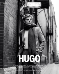 Black and white portrait of a young man wearing a grey suit and scarf standing in an alleyway for The Perfect Man Magazine