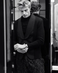 Black and white portrait of a blonde male model adjusting his watch while wearing a black tuxedo jacket and textured trousers.