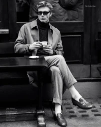 Man wearing sunglasses and a coat seated at an outdoor café table holding a small white coffee cup
