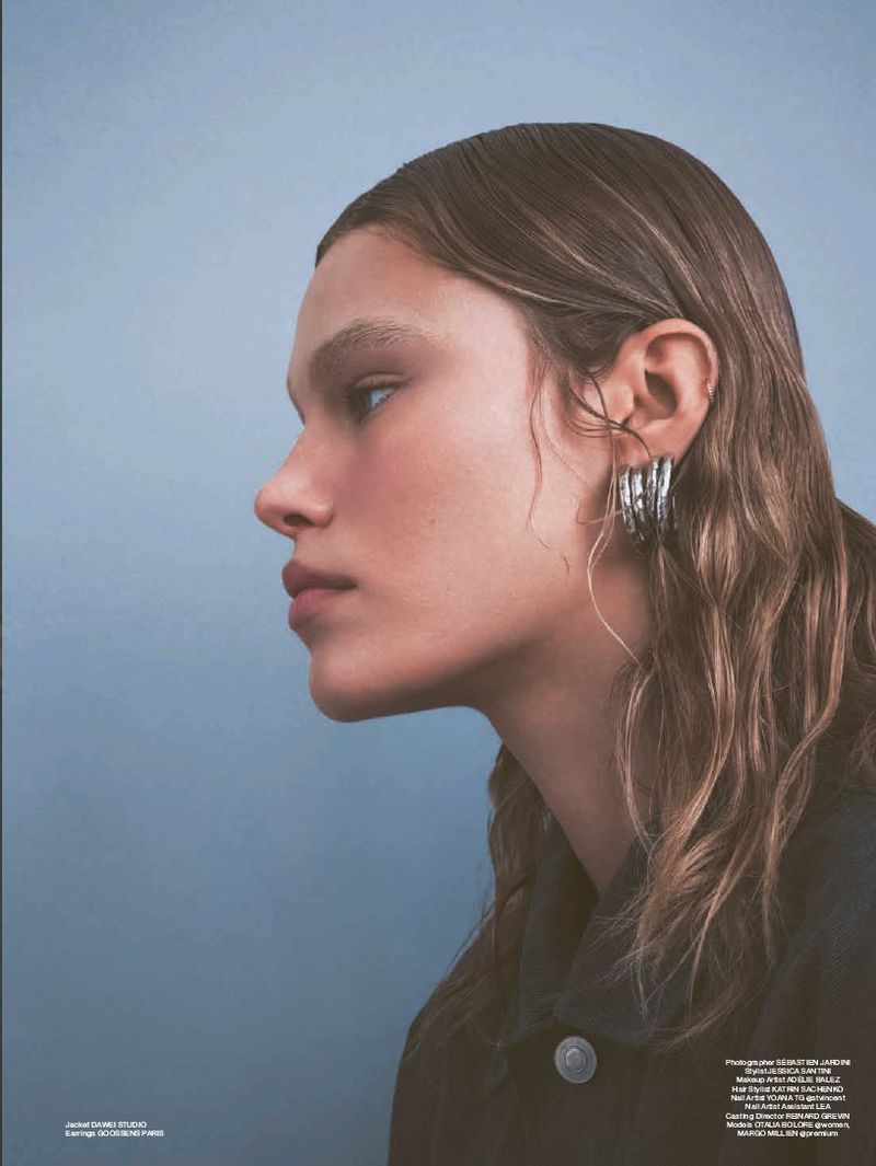 Profile shot of a woman with wavy brown hair wearing silver hoop earrings against a blue background in a fashion editorial.