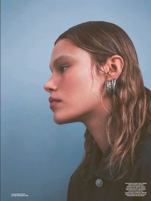 Side profile portrait of a woman with wet-look hair wearing chunky silver statement earrings and a dark jacket against a light blue backdrop.