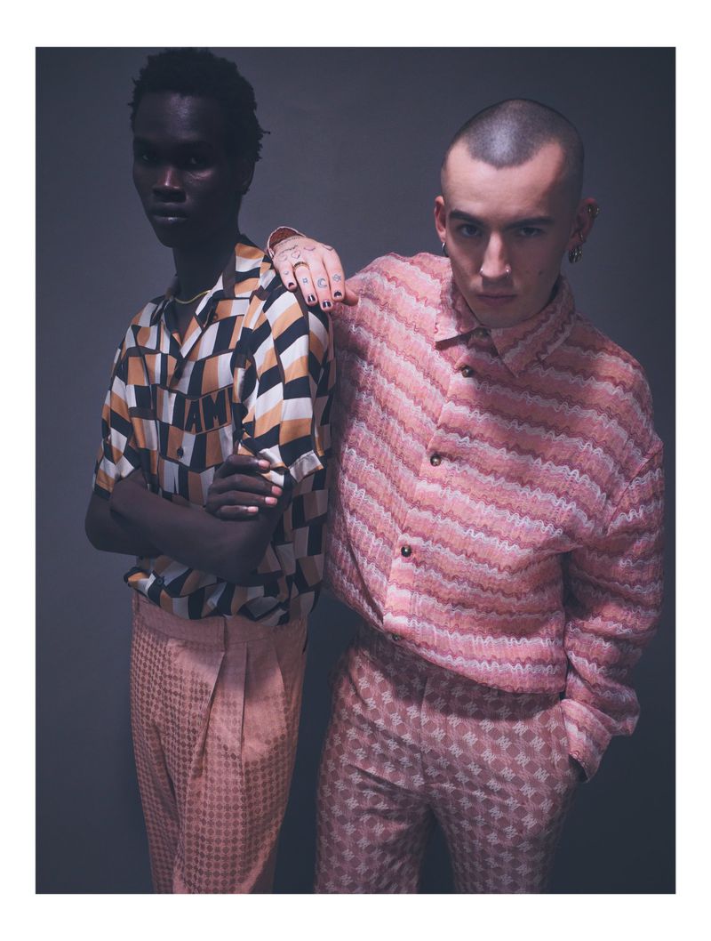 Two fashion models pose indoors against a gray background, wearing patterned shirts and pants. A dark-skinned model rests a hand on another model.