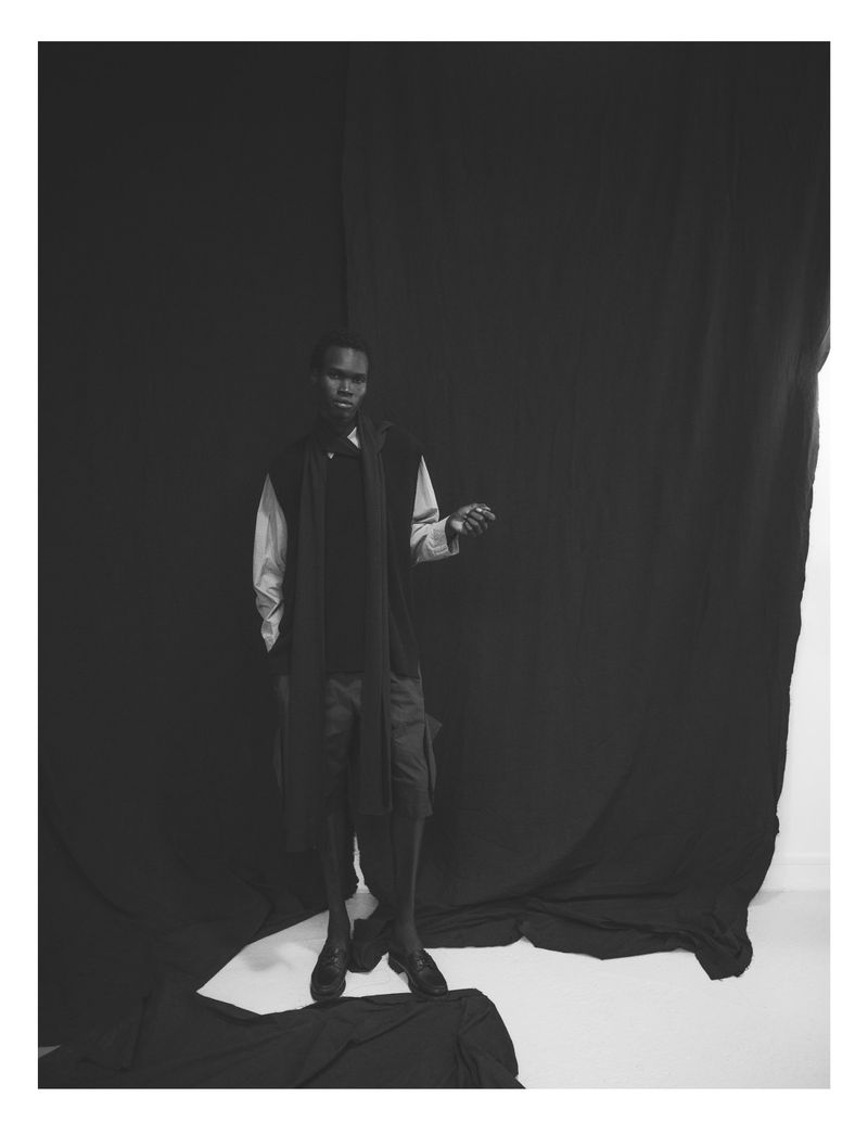 Black and white portrait of a Black male model posing in an indoor studio with a black draped backdrop, wearing a light shirt and dark vest.