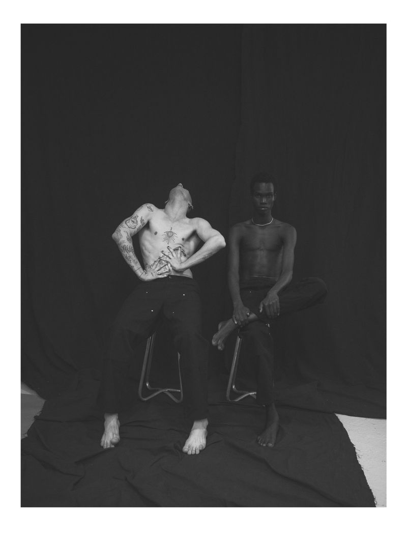 Two shirtless men sit on metal stools in a monochromatic photo studio setting, tattoos visible on one man, against a draped black backdrop.