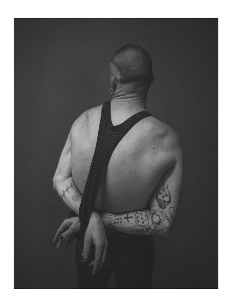 A black-and-white back shot of an adult with tattoos, hands clasped behind, wearing a knitted halter top against a gray backdrop.