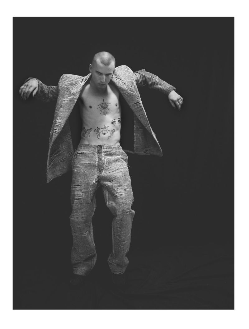 Monochrome full shot of an adult male with tattoos wearing an open jacket and jeans, in a poised position against a dark background.