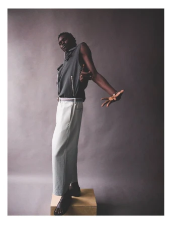 Adult male poses in a sleeveless gray top and light-colored pants, standing on a wooden box against a muted gray backdrop in a studio.