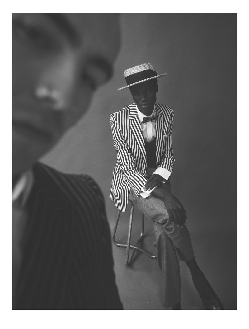 Black and white portrait of African male in striped jacket and boater hat, seated on metal stool in photo studio, with blurred person in foreground.