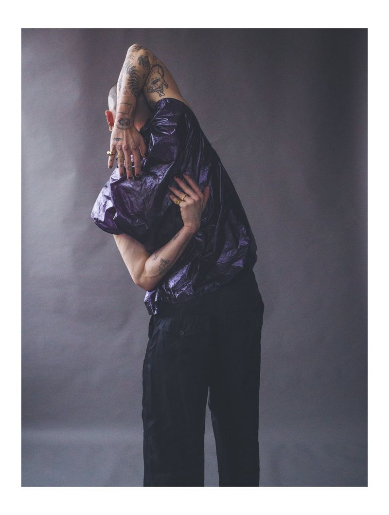 A person with tattoos strikes a pose in a shiny purple top and black pants against a grey background in a studio shot.