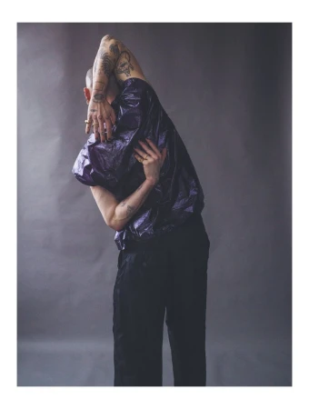 A person with tattoos strikes a pose in a shiny purple top and black pants against a grey background in a studio shot.