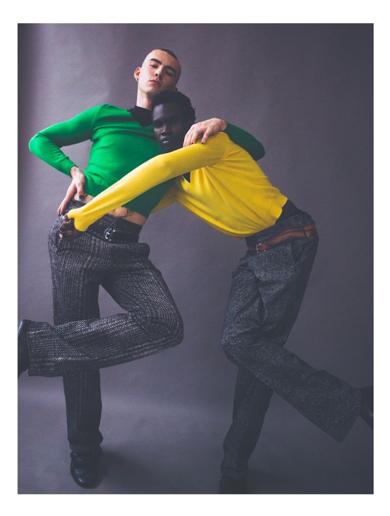Two male fashion models in vibrant sweaters and patterned pants pose in a studio with soft lighting against a plain backdrop.