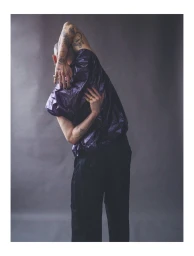 Person with tattooed arms wearing a metallic purple shirt and dark trousers against a grey backdrop