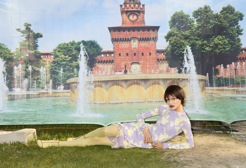 A woman lies on grass in a lavender and yellow dress in front of a backdrop of a castle, fountain, and trees.