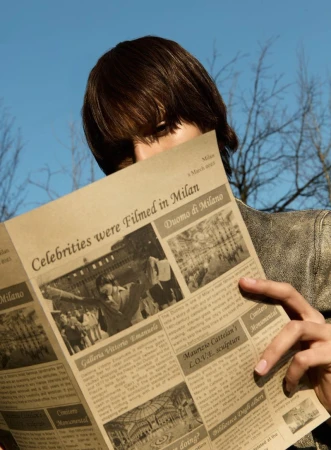 Person reading a newspaper with headlines about celebrities and Milan landmarks against a blue sky and bare tree branches.