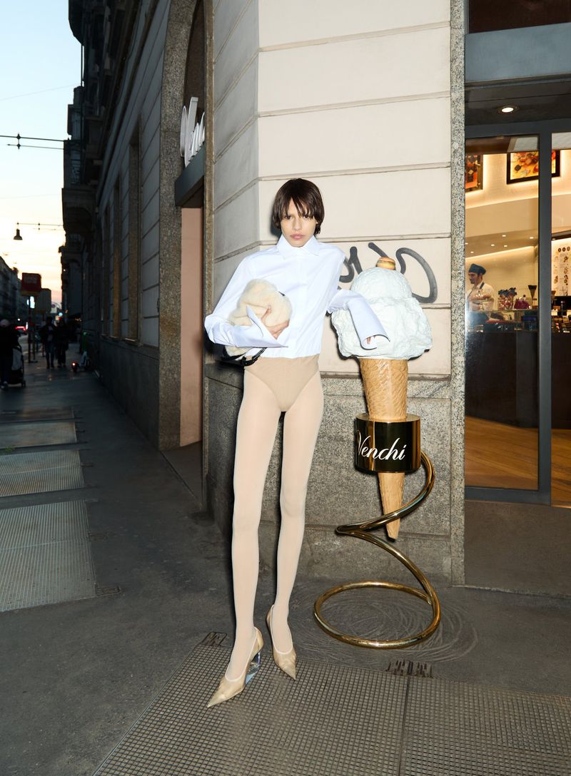 Woman in white shirt and beige tights poses next to an oversized ice cream cone prop outside of a shop in an urban environment.