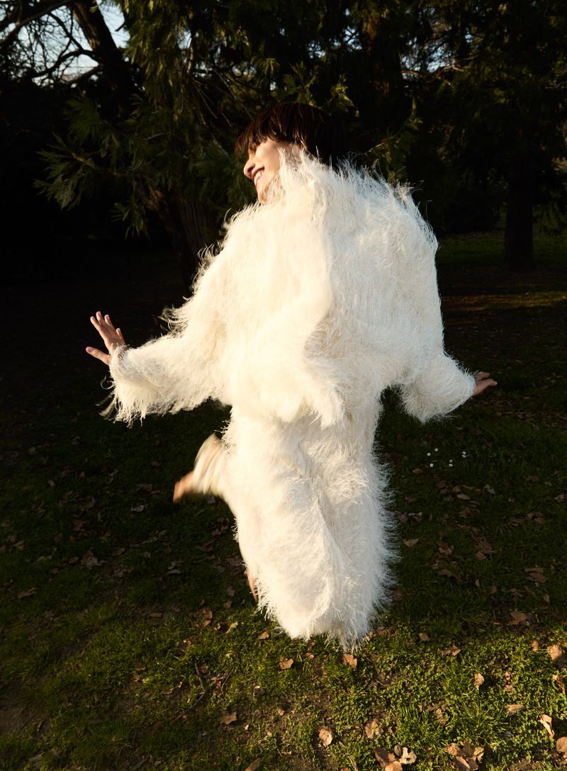 A woman jumps in a grassy park wearing a furry white coat, with trees and a bright sky creating a dynamic outdoor fashion moment.