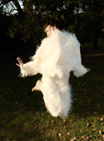 A woman jumps in a grassy park wearing a furry white coat, with trees and a bright sky creating a dynamic outdoor fashion moment.