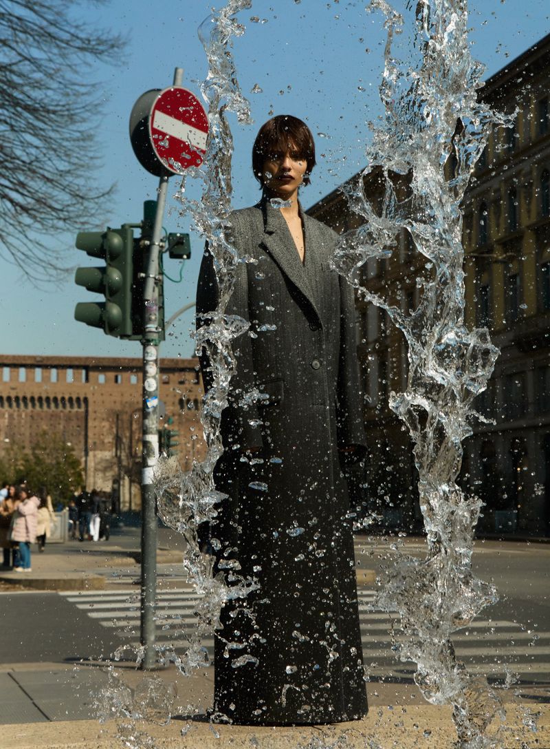 Woman in grey coat stands behind water fountain on city street with traffic light and European architecture in background on sunny day.