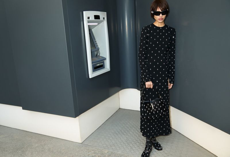 Adult woman in patterned dress standing near ATM indoors, wearing sunglasses and holding a black purse, fashion lifestyle.