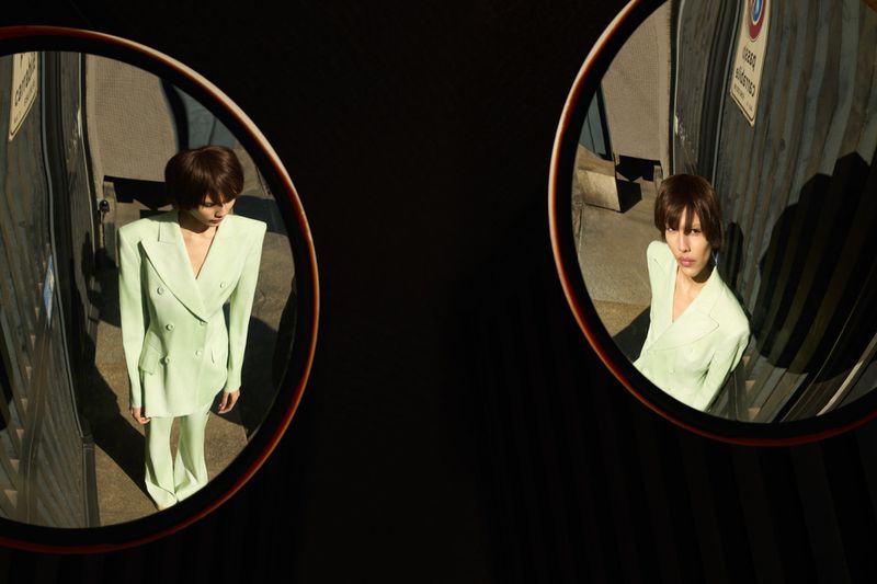 Woman in a mint green suit reflected in round mirrors against an urban backdrop, creating a symmetrical and stylized fashion image.