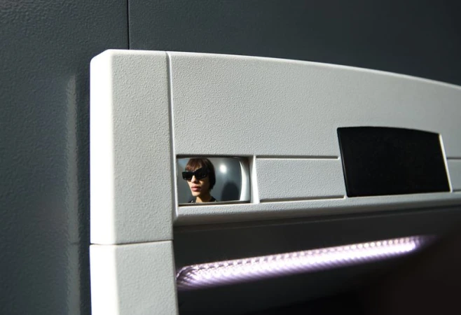 An adult looking out from a square opening in a white wall wears sunglasses, illuminated by a light strip below in an indoor scene.