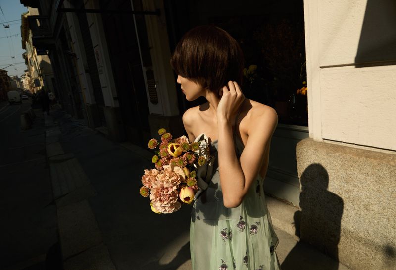 Fashion photo of an adult woman with a bouquet of flowers standing on an urban street, lit by sunlight with building shadows.