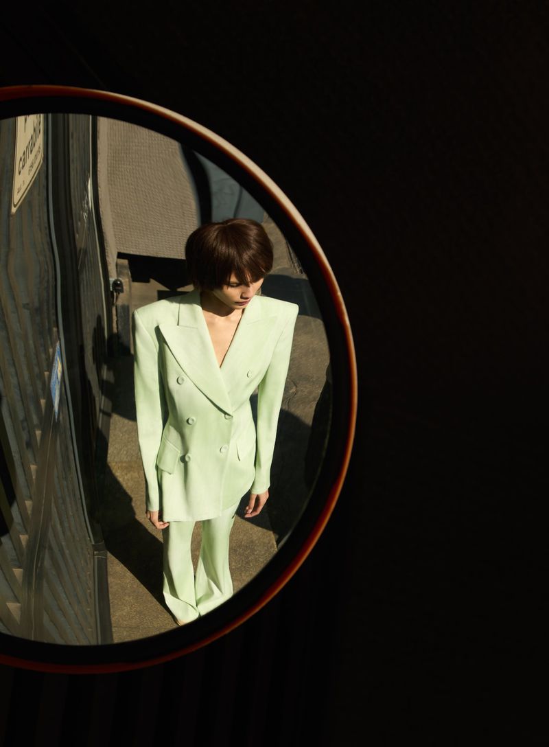 Mirror reflects a person in a light green pantsuit standing outdoors on an overcast day in sunlight against a dark background.