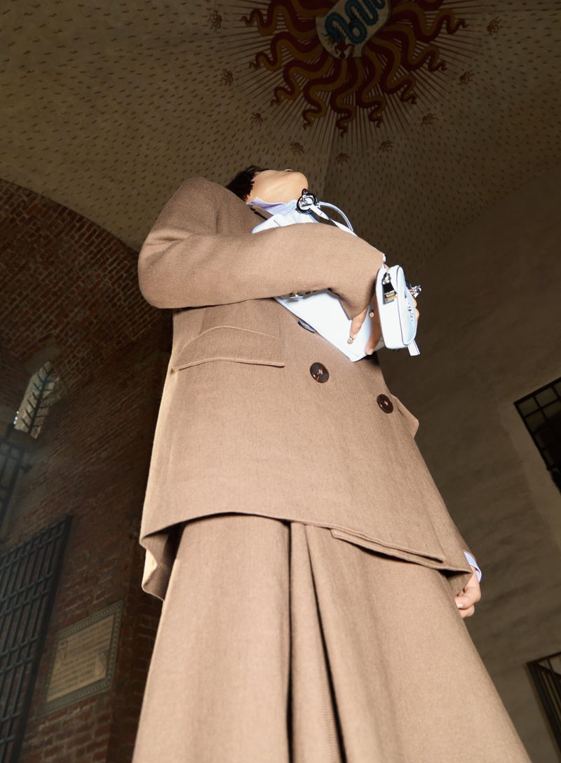 Woman in a brown suit and white bag in front of a brick building looking upwards. Low angle and neutral expression.
