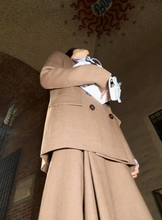 Woman in a brown suit and white bag in front of a brick building looking upwards. Low angle and neutral expression.